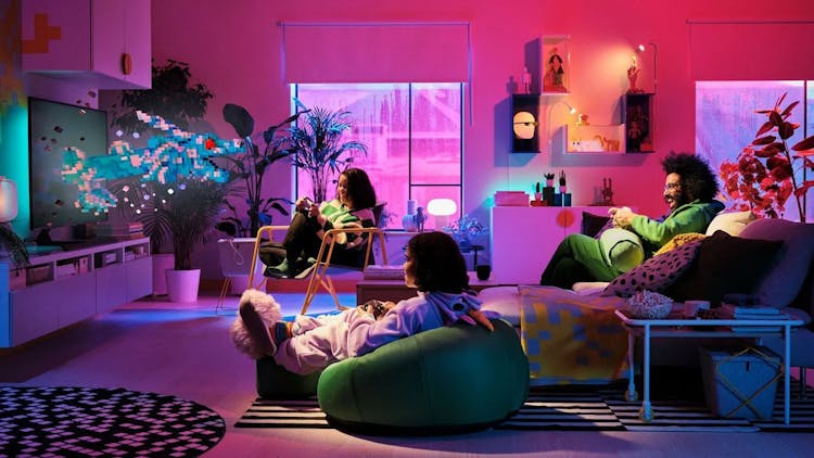 Three people gaming in a cozy, colorful living room with vibrant lighting and pixelated art on the wall.